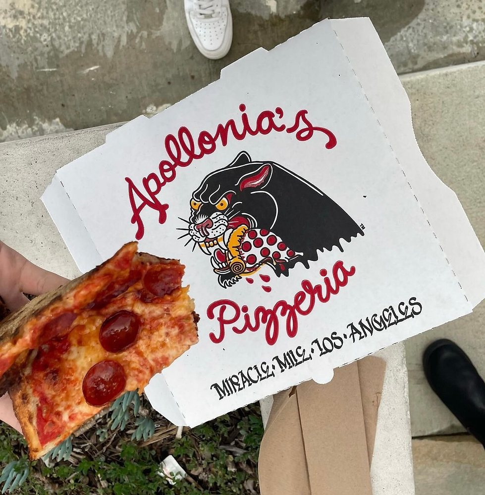 Hand holding a folded pepperoni pizza slice in front of an Apollonia's Pizzeria delivery box printed with a black panther devouring a slice, on a Miracle Mile sidewalk