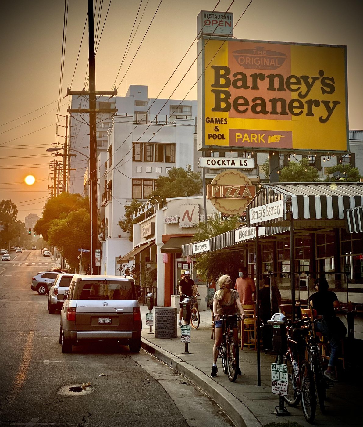 Barney's Beanery's yellow rooftop sign reading Games & Pool and Park above its striped awning on Santa Monica Boulevard at sunset, with cyclists on the sidewalk